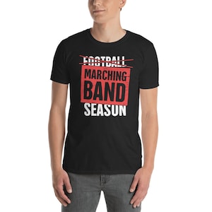 Può includere: Maglietta nera con una grafica rossa e bianca che dice "Football" con una linea barrata e "Marching Band Season" sotto.