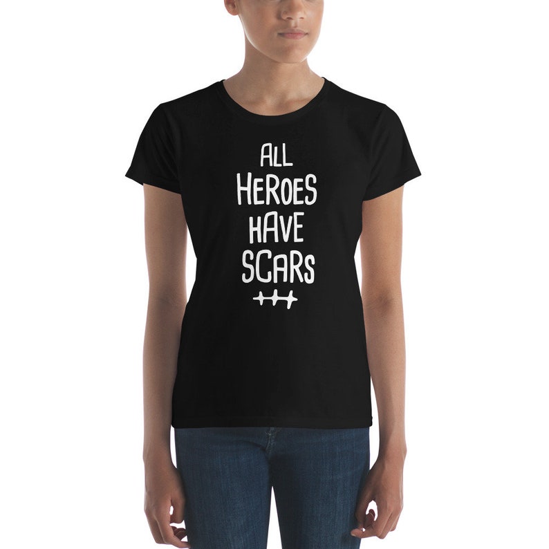 K&ouml;nnte beinhalten: Schwarzes Damen-T-Shirt mit wei&szlig;em Text, der "All Heroes Have Scars" lautet, mit drei Pluszeichen darunter.
