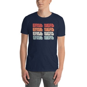 Può includere: Una t-shirt blu navy con il testo "Band Nerd" ripetuto quattro volte in un font in stile vintage con un gradiente di rosso, arancione, giallo e blu.
