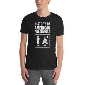 Op de afbeelding: Zwart T-shirt met witte tekst die "Geschiedenis van Amerikaanse presidenten" luidt. Het shirt heeft een afbeelding van een stokfiguur die de eerste 44 presidenten voorstelt en een clownfiguur die de 45e president voorstelt.