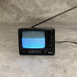Puede incluir: Un pequeño televisor negro vintage con una antena plateada extendida hacia arriba. La pantalla muestra estática con ruido azul y blanco. El televisor tiene un dial y la marca "SEIKO" es visible.