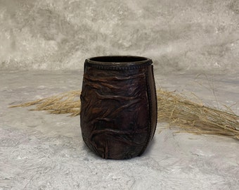 May include: A brown leather cup with a textured surface. The cup is made of stitched leather panels and has a slightly rounded shape.