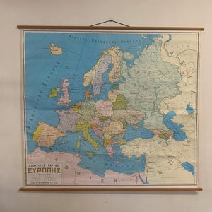 May include: A large wall-mounted political map of Europe printed on paper with wooden dowels at the top and bottom for hanging. The map features various countries in muted pastel colours, blue oceans, and Greek text including the title ΕΥΡΩΠΗΣ.