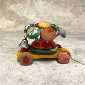 Może przedstawiać: Drewniana zabawka do ciągnięcia z epoki z żółtą podstawą i czerwonymi, zielonymi i żółtymi kołami. Zabawka ma dzwonek i obracający się wierzchołek z kolorowymi, malowanymi drewnianymi gałkami.