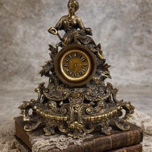 Può includere: Un orologio antico in ottone con una figura femminile seduta in cima. Il quadrante dell'orologio presenta numeri romani ed è circondato da dettagli ornati. L'orologio è posto sopra due libri antichi.