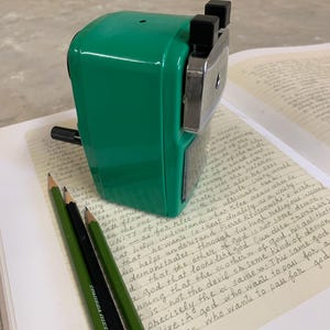 K&ouml;nnte beinhalten: Ein gr&uuml;ner Bleistiftanspitzer mit schwarzen Akzenten liegt auf einem Notizbuch mit handschriftlichem Text. Drei Bleistifte, zwei gr&uuml;ne und einer schwarz mit einer orangefarbenen und roten Spitze, liegen daneben. Das Bild hat eine Vintage-&Auml;sthetik.