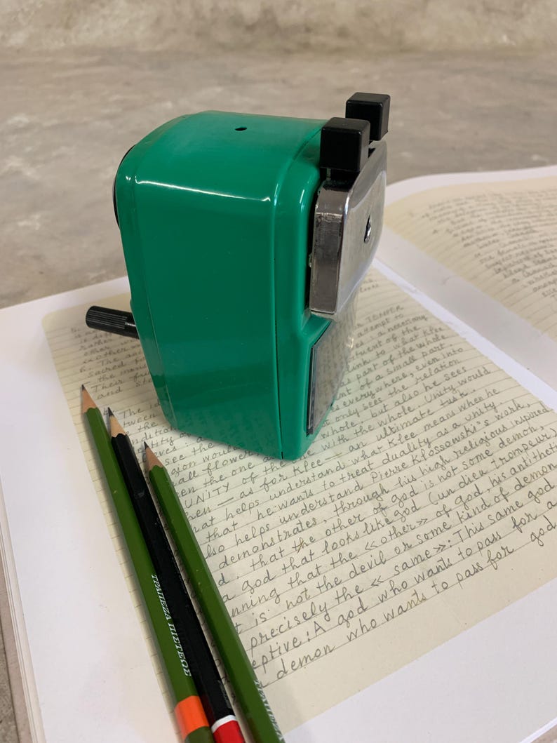 K&ouml;nnte beinhalten: Ein gr&uuml;ner Bleistiftanspitzer mit schwarzen Akzenten liegt auf einem Notizbuch mit handschriftlichem Text. Drei Bleistifte, zwei gr&uuml;ne und einer schwarz mit einer orangefarbenen und roten Spitze, liegen daneben. Das Bild hat eine Vintage-&Auml;sthetik.