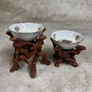 May include: Two white porcelain bowls with floral designs, each sitting on a wooden stand with intricate carvings. The stands are stacked on top of each other.
