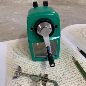 K&ouml;nnte beinhalten: Ein gr&uuml;ner manueller Bleistiftanspitzer mit silberner Kurbel und schwarzen Akzenten. Der Anspitzer liegt auf einem Notizbuch mit handschriftlichem Text, zusammen mit Bleistiften und einer Klemme.