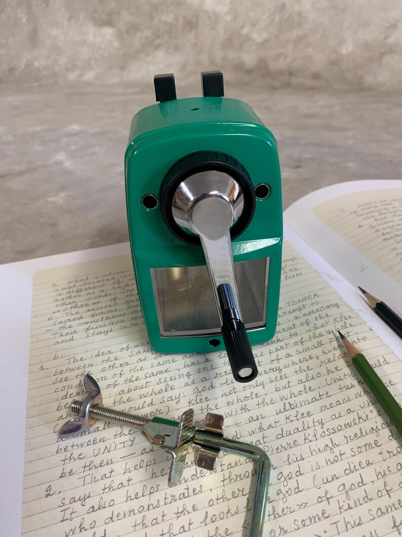 K&ouml;nnte beinhalten: Ein gr&uuml;ner manueller Bleistiftanspitzer mit silberner Kurbel und schwarzen Akzenten. Der Anspitzer liegt auf einem Notizbuch mit handschriftlichem Text, zusammen mit Bleistiften und einer Klemme.