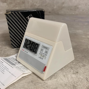May include: A cream-coloured digital alarm clock displaying 12:08. The clock features a red accent and is accompanied by its original box and instruction manual. The box is black with white diagonal stripes. The clock's dimensions are in centimetres.