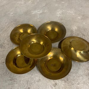 May include: Six small, antique brass bowls. Each bowl features a rounded, concave centre and a decorative, embossed rim. The brass has a warm, golden hue, with slight variations in colour and patina, suggesting age and use.