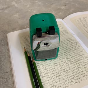 K&ouml;nnte beinhalten: Ein gr&uuml;ner Bleistiftanspitzer mit einer silbernen Klinge und zwei schwarzen Kn&ouml;pfen liegt auf einem Notizbuch mit handschriftlichem Text. Drei Bleistifte, zwei gr&uuml;ne und einer schwarzer, liegen neben dem Anspitzer. Die Bleistifte haben orange und rote Streifen.