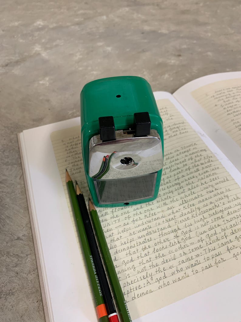K&ouml;nnte beinhalten: Ein gr&uuml;ner Bleistiftanspitzer mit einer silbernen Klinge und zwei schwarzen Kn&ouml;pfen liegt auf einem Notizbuch mit handschriftlichem Text. Drei Bleistifte, zwei gr&uuml;ne und einer schwarzer, liegen neben dem Anspitzer. Die Bleistifte haben orange und rote Streifen.
