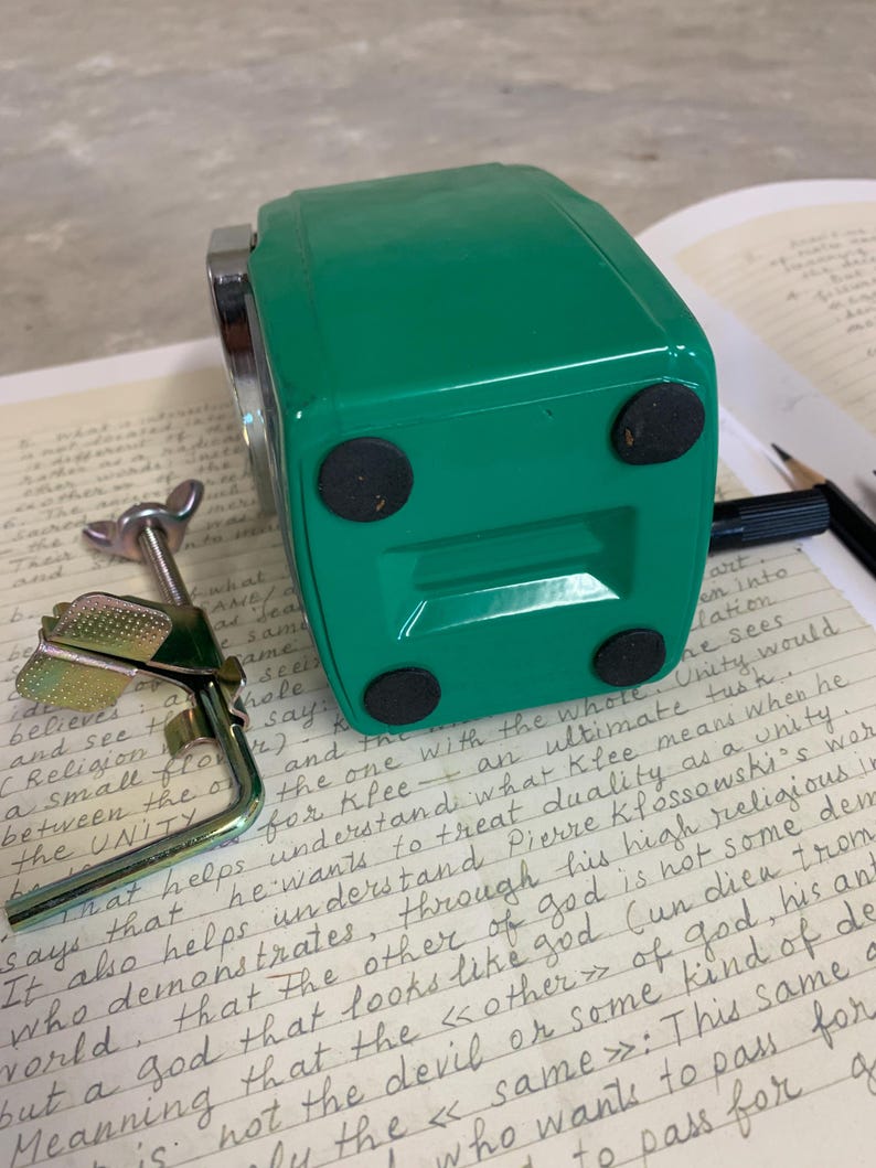 K&ouml;nnte beinhalten: Ein gr&uuml;ner, rechteckiger Anspitzer mit schwarzen Gummif&uuml;&szlig;en. Der Anspitzer liegt auf einem Blatt linierten Papiers mit handschriftlichen Notizen und einem Bleistift.