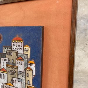 May include: Framed ceramic tile art depicting a stylized village scene with white buildings, red and yellow accents, and a blue sky. The artwork is set within a wooden frame and a peach-colored matting.