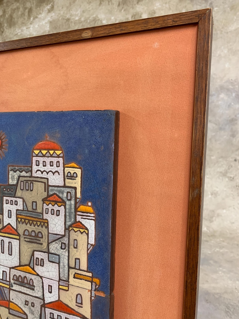 May include: Framed ceramic tile art depicting a stylized village scene with white buildings, red and yellow accents, and a blue sky. The artwork is set within a wooden frame and a peach-colored matting.