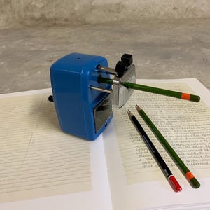 Peut inclure: Un taille-crayon bleu avec une lame argentée taille un crayon vert. Deux crayons supplémentaires, un noir et un vert, reposent sur une page de notes manuscrites. Le taille-crayon est sur une surface blanche.