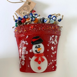 Könnte beinhalten: Roter Glasornament in Topfform mit Schneemann-Design. Der Schneemann hat einen schwarzen Hut, eine orangefarbene Nase und einen roten Schal. Die Oberseite des Topfes ist mit Perlen gefüllt und trägt ein Etikett mit der Aufschrift "Hand Made for You".