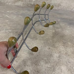 Op de afbeelding: Een chromen metalen kapstok met acht gebogen haken. Elke haak heeft een druppelvormige, olijfgroene en bruin gespikkelde knop. De kapstok is ontworpen om aan de muur te worden bevestigd.