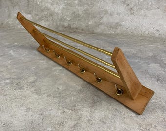 May include: Wooden wall-mounted coat rack with brass hooks and bars. The rack has a light brown finish and a mid-century modern design. Six brass hooks are aligned along the bottom, with two brass bars above.