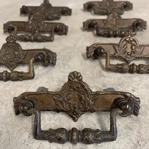 May include: A collection of antique bronze-toned drawer pulls. Each pull features an ornate, detailed design with a central decorative element and curved handles. The pulls are arranged on a neutral surface, showcasing their vintage aesthetic.