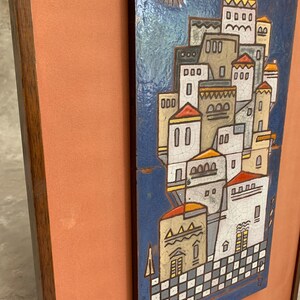 May include: A rectangular wall plaque featuring a ceramic tile with a blue background and a stylized village scene. The scene includes white and tan buildings with red roofs, a red sun, and a checkered base, all framed in a dark wood border.
