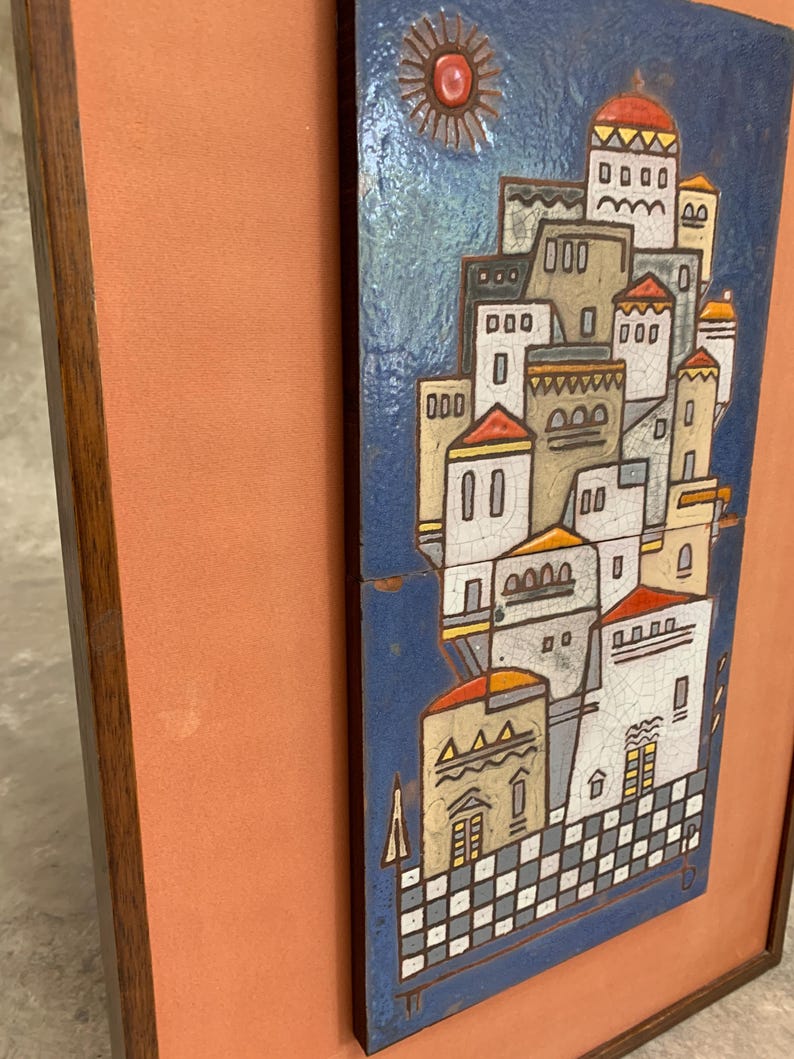 May include: A rectangular wall plaque featuring a ceramic tile with a blue background and a stylized village scene. The scene includes white and tan buildings with red roofs, a red sun, and a checkered base, all framed in a dark wood border.