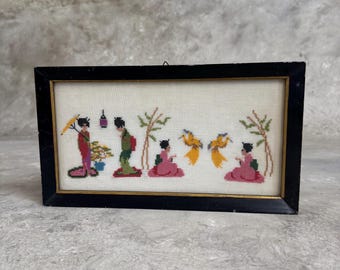 May include: A framed cross-stitch embroidery depicting a scene of Japanese women in traditional clothing with birds and bamboo plants. The embroidery is done in shades of pink, green, yellow, and black.