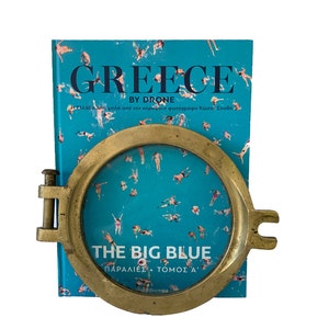 K&ouml;nnte beinhalten: Ein Messing-Bullaugenrahmen umrahmt ein Buch mit dem Titel "GREECE BY DRONE" mit einem t&uuml;rkisfarbenen Einband mit Schwimmern. Das Buch zeigt auch den Titel "THE BIG BLUE". Das Bullauge hat ein Scharnierdesign und steht vor einem wei&szlig;en Hintergrund.