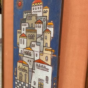 May include: A rectangular ceramic wall plaque featuring a blue background with a stylized village scene of white buildings with orange roofs. The artwork has a visible crackle glaze finish and is framed in a dark wood border with a peach-colored mat.