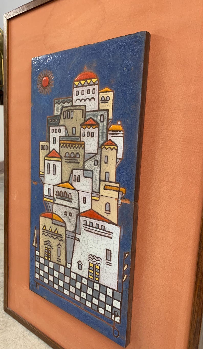 May include: A rectangular ceramic wall plaque featuring a blue background with a stylized village scene of white buildings with orange roofs. The artwork has a visible crackle glaze finish and is framed in a dark wood border with a peach-colored mat.