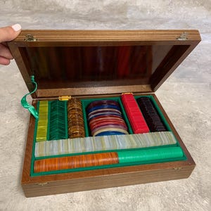 Könnte beinhalten: Eine Holzschatulle mit Klappdeckel, die eine Sammlung bunter Spielchips enthält. Das Innere ist mit grünem Filz ausgekleidet und verfügt über organisierte Fächer, die Stapel von rechteckigen und runden Chips in Rot, Grün, Orange, Braun und Blau enthalten.