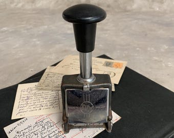 May include: Vintage metal number stamp with a black handle and a silver base. The stamp is labeled "Made in England." The stamp is resting on a stack of old letters and postcards, creating a nostalgic scene.