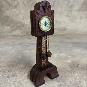 May include: A miniature wooden grandfather clock with a gold-colored face and a pendulum. The clock is carved with intricate details and has a dark brown finish.