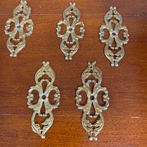 May include: Five ornate, gold-coloured metal decorative hardware pieces. Each piece features a symmetrical, scrolling design with a central opening. The hardware is likely for furniture or decorative applications.