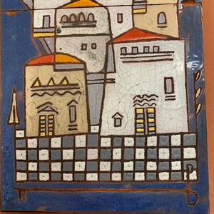 May include: A rectangular ceramic tile featuring a colorful, abstract depiction of a village. The artwork showcases buildings with red, yellow, and beige roofs, set against a blue background. The tile has a cracked glaze effect, adding texture.