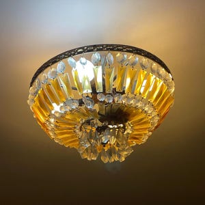 以下が含まれることがあります： 円形のデザインのヴィンテージシーリングライト。この照明器具は、暗い金属フレームが特徴で、透明と琥珀色のクリスタルアクセントで飾られています。クリスタルは層状に配置されており、装飾的でエレガントな外観を作り出しています。