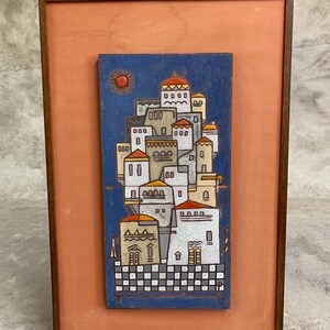 May include: A framed ceramic wall plaque featuring a blue background with a cluster of white and beige buildings, orange roofs, and a sun. The artwork is set against a terracotta-colored mat within a thin, dark brown wooden frame.