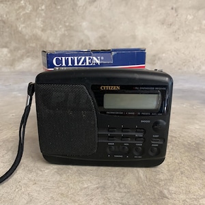 Puede incluir: Radio portátil Citizen negra con pantalla digital y varios botones. La radio incluye el texto "CITIZEN" y "PLL SYNTHESIZED RECEIVER". Una pequeña caja con el logotipo de Citizen está detrás de la radio.