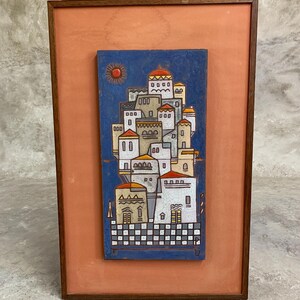 May include: A framed artwork featuring a stylized village scene. The artwork depicts white buildings with orange and yellow roofs against a blue background. A red sun is in the upper left corner. The frame is a dark brown color.