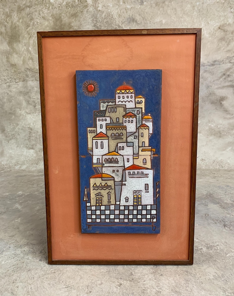 May include: A framed artwork featuring a stylized village scene. The artwork depicts white buildings with orange and yellow roofs against a blue background. A red sun is in the upper left corner. The frame is a dark brown color.