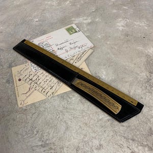 Op de afbeelding: Een vintage liniaal met een decoratief ontwerp, tot 30 cm, is gehuisvest in een zwart leren etui. De liniaal is gemaakt van messing en heeft een decoratief patroon. Oude ansichtkaarten staan op de achtergrond.