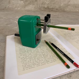 K&ouml;nnte beinhalten: Ein gr&uuml;ner Bleistiftanspitzer mit einem eingesetzten Bleistift, der auf einem offenen Buch mit handschriftlichem Text liegt. Zwei zus&auml;tzliche Bleistifte, einer gr&uuml;n und einer schwarz, liegen neben dem Anspitzer. Die Bleistifte haben orange und rote Akzente.