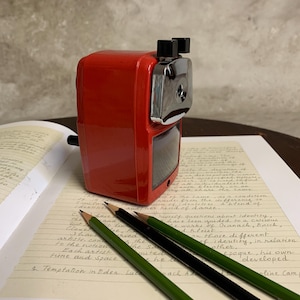 Pode incluir: Um apontador de lápis vermelho, vintage, com uma carcaça de lâmina prateada repousa sobre uma página de notas manuscritas. Três lápis, verdes e pretos, estão dispostos em frente ao apontador. A cena evoca nostalgia.