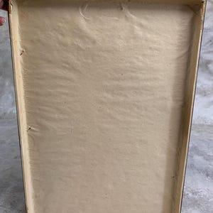 May include: The back of a rectangular picture frame featuring a light brown paper backing secured within a dark wooden border. A small metal triangular hanging hook is attached to the top center of the frame's wooden edge.