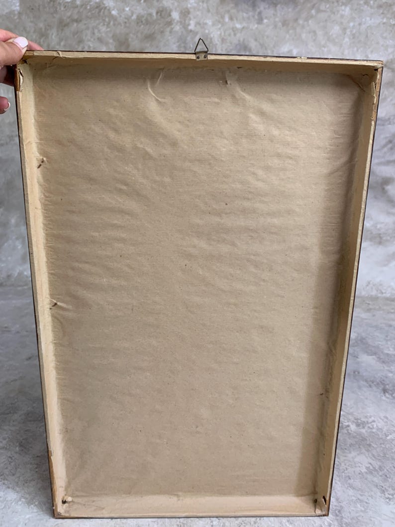 May include: The back of a rectangular picture frame featuring a light brown paper backing secured within a dark wooden border. A small metal triangular hanging hook is attached to the top center of the frame's wooden edge.