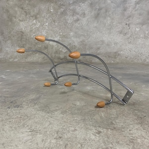 Può includere: Un attaccapanni da parete in metallo argentato con sei pomelli in legno. L'attaccapanni ha un design curvo con una piastra di montaggio rettangolare. I pomelli sono marrone chiaro e di forma ovale.