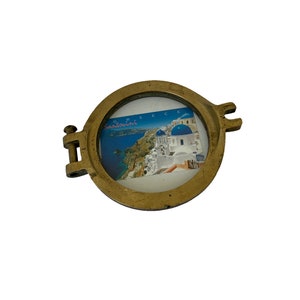 K&ouml;nnte beinhalten: Ein runder, messingfarbener Bilderrahmen mit einer Scharnierseite und zwei dekorativen Laschen. Der Rahmen enth&auml;lt eine Postkarte von Santorini, Griechenland, mit blau-bedachten Geb&auml;uden und dem Wort "Griechenland". Der Rahmen befindet sich auf wei&szlig;em Hintergrund.