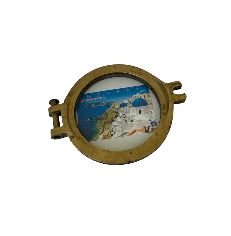 K&ouml;nnte beinhalten: Ein runder, messingfarbener Bilderrahmen mit einer Scharnierseite und zwei dekorativen Laschen. Der Rahmen enth&auml;lt eine Postkarte von Santorini, Griechenland, mit blau-bedachten Geb&auml;uden und dem Wort "Griechenland". Der Rahmen befindet sich auf wei&szlig;em Hintergrund.